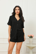 A woman standing in a room wearing a black short pyjama set with a button-up front and drawstring waist.